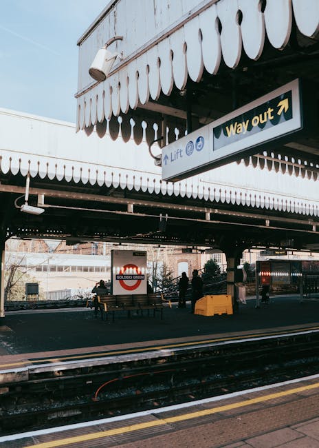 An exterior view of a train station platform with a covered, arched metal roof structure and brick building facades in the background. The platform has tactile paving along its edge and is marked with